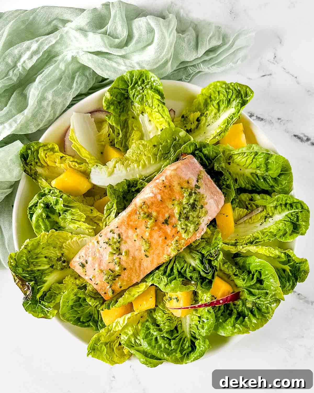 A fillet of salmon sits on a bed of little gem lettuce, mango, red onion, and jalapeno vinaigrette on a white marble counter.
