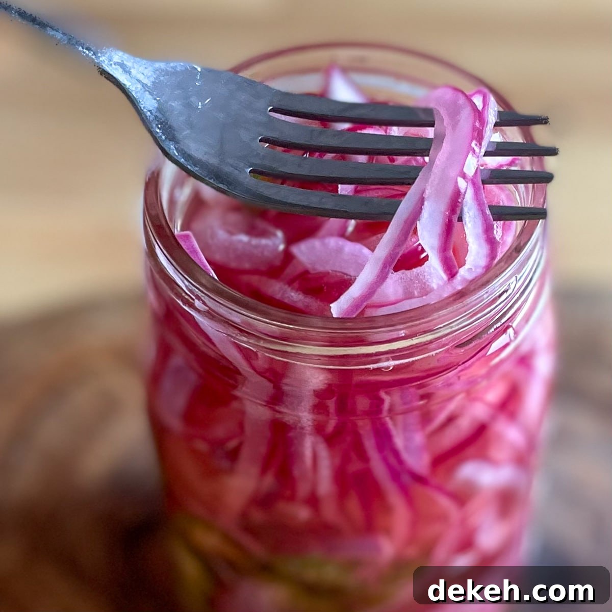 Sliced red spicy pickled onions are lifted out of a glass Mason jar with a silver fork.