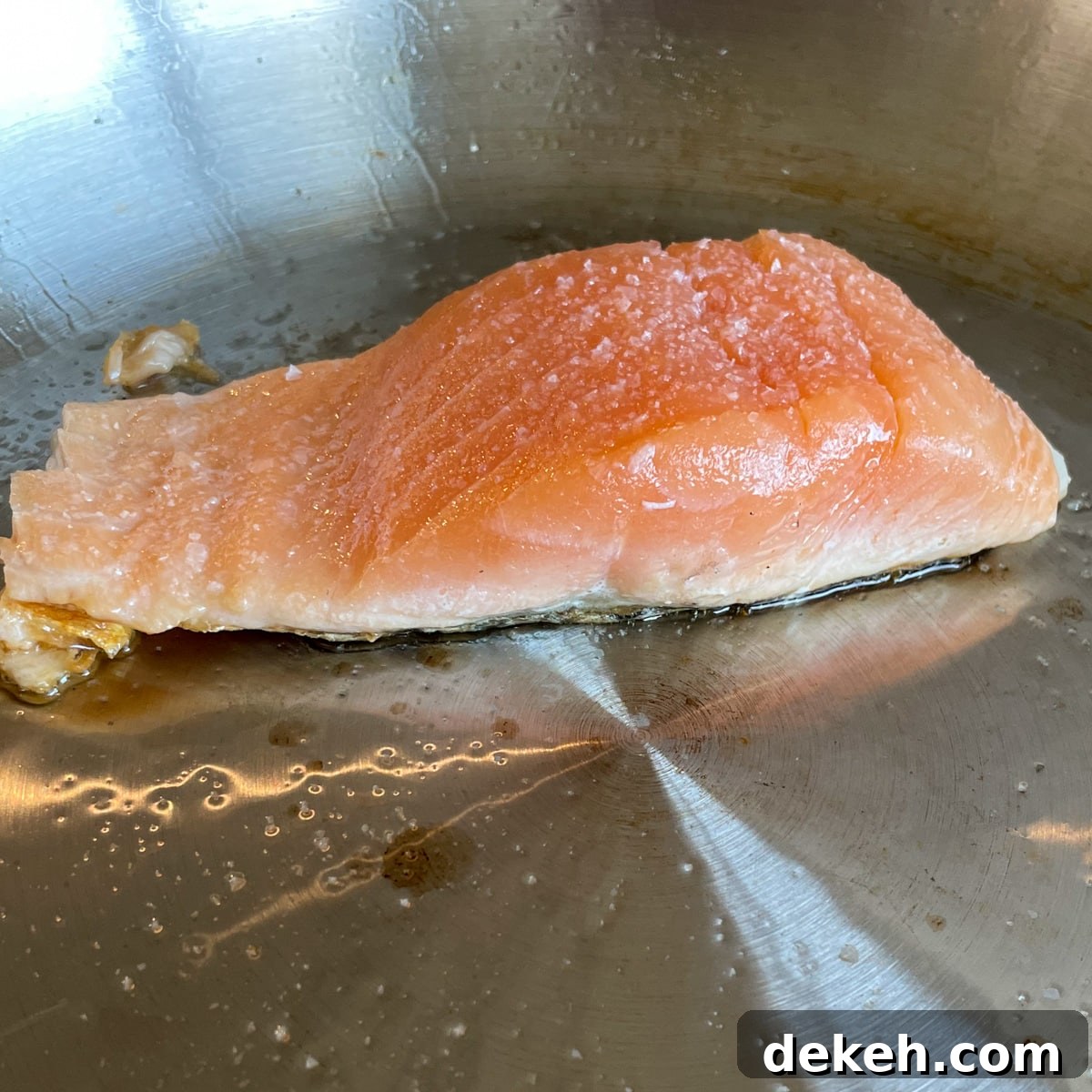 A fillet of salmon is cooking in a stainless steel pan with olive oil and salt. The salmon changes color halfway through, showing it is half cooked.