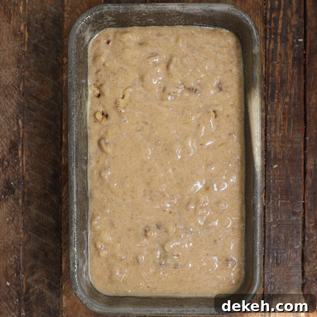 A metal loaf pan filled with unbaked Banana Bread batter, its lumpy mixture with visible chunks on display, sits invitingly on a rustic wooden surface.
