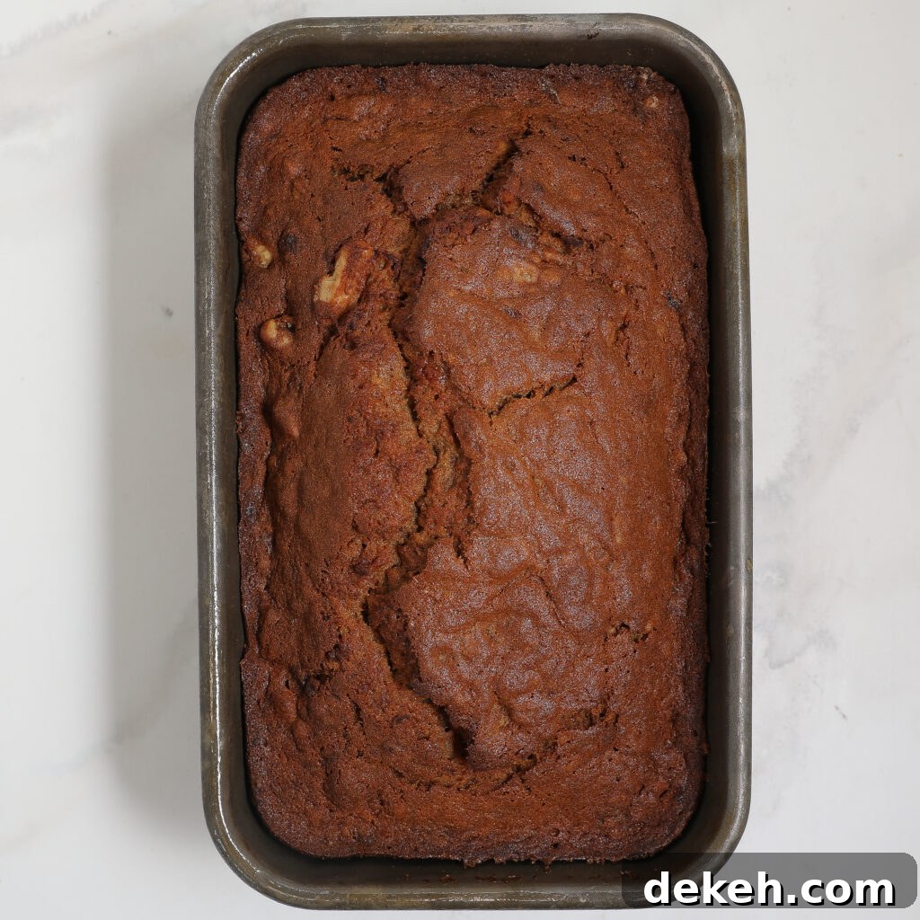 A loaf of Banana Bread with a golden-brown, cracked top sits in a rectangular metal baking pan on a white surface.