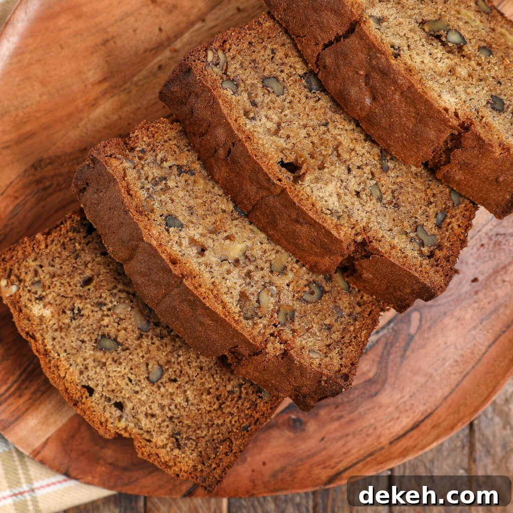 Four slices of banana bread with visible walnut pieces are arranged on a wooden plate, set on a rustic wooden surface.
