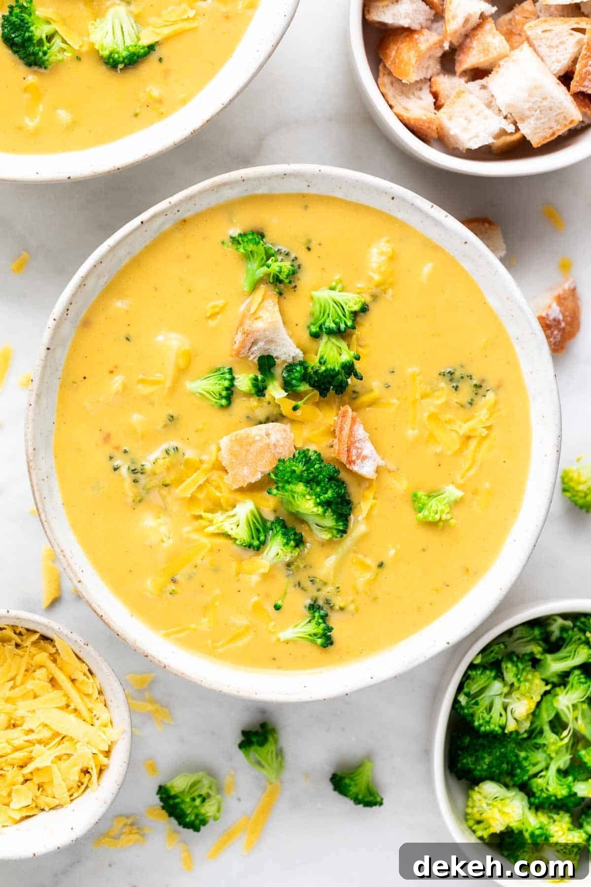 Vegan broccoli cheddar soup in a bowl garnished with bread and cheddar cheese, with toppings around.