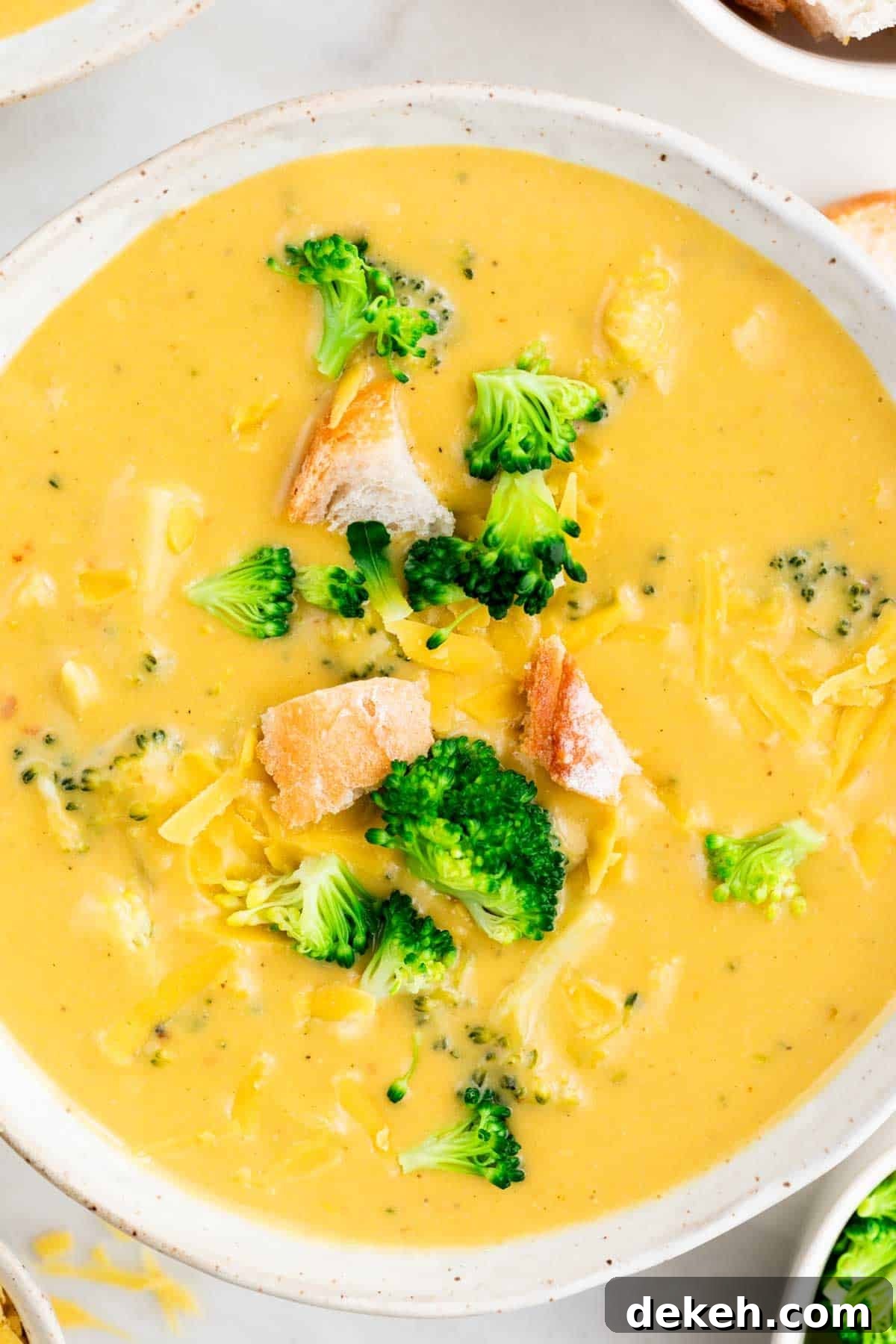 Vegan broccoli cheddar soup in a bowl topped with bread and cheese, surrounded by additional toppings.