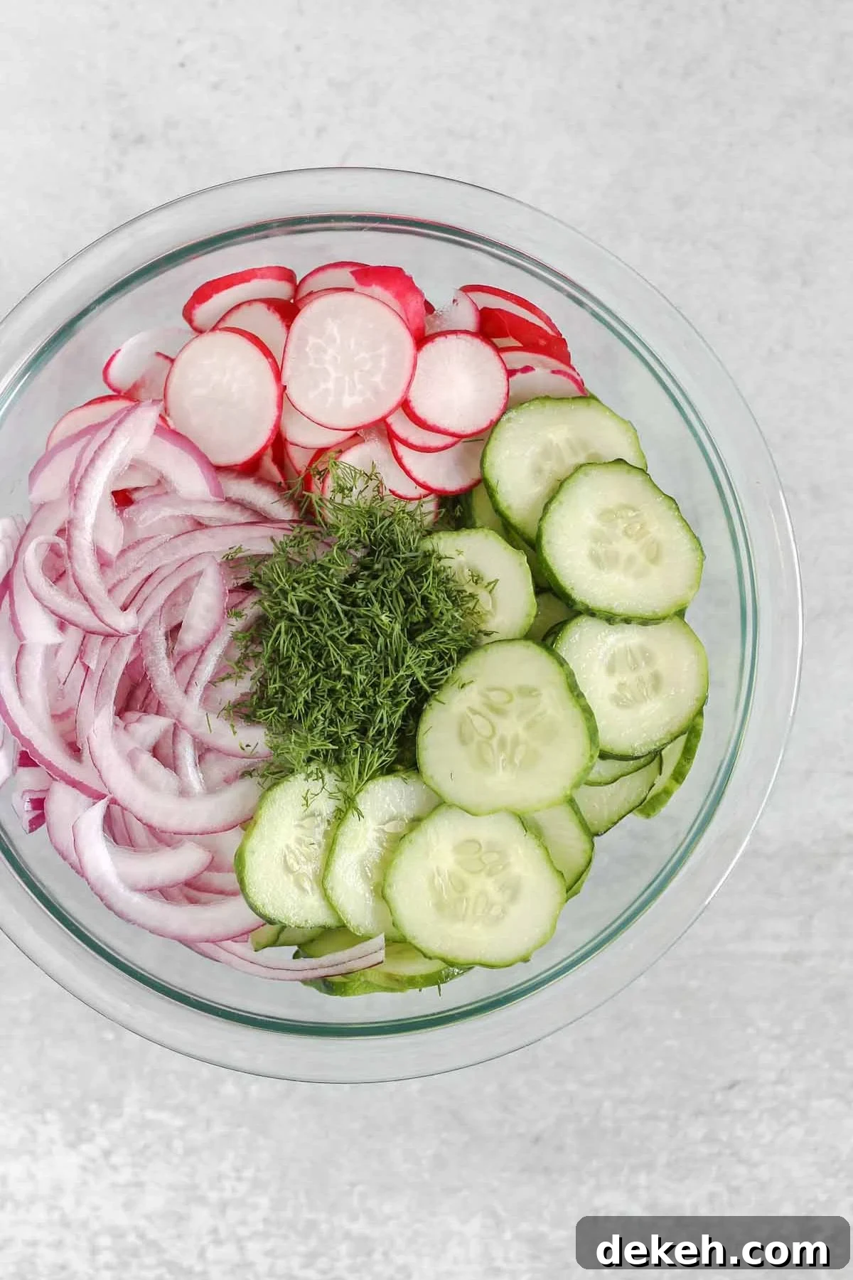 Slice cucumbers, sliced radishes, sliced red onion, and fresh dill in a glass mixing bowl.