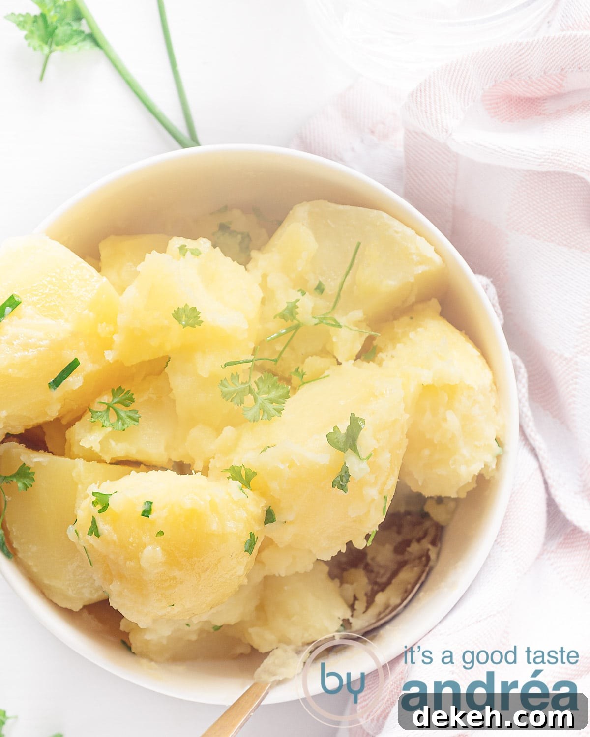 A white bowl filled with potatoes cooked with chives and parsley on a white background with a pink-and-white tea towel