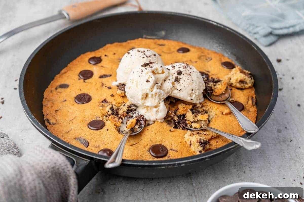Overhead shot of a warm chocolate chip skillet cookie in a black skillet, topped with scoops of vanilla ice cream and three spoons.
