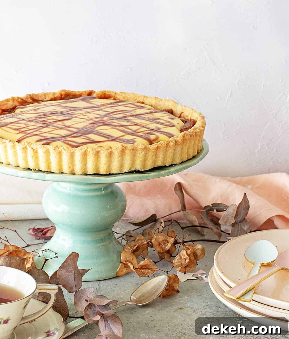 Whole lemon tart on a green cake stand. Stack of pink plates, dried leaves, a grey surface. 