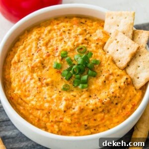 A bowl of vegan roasted red pepper dip garnished with chopped green onion and with four crackers in it.