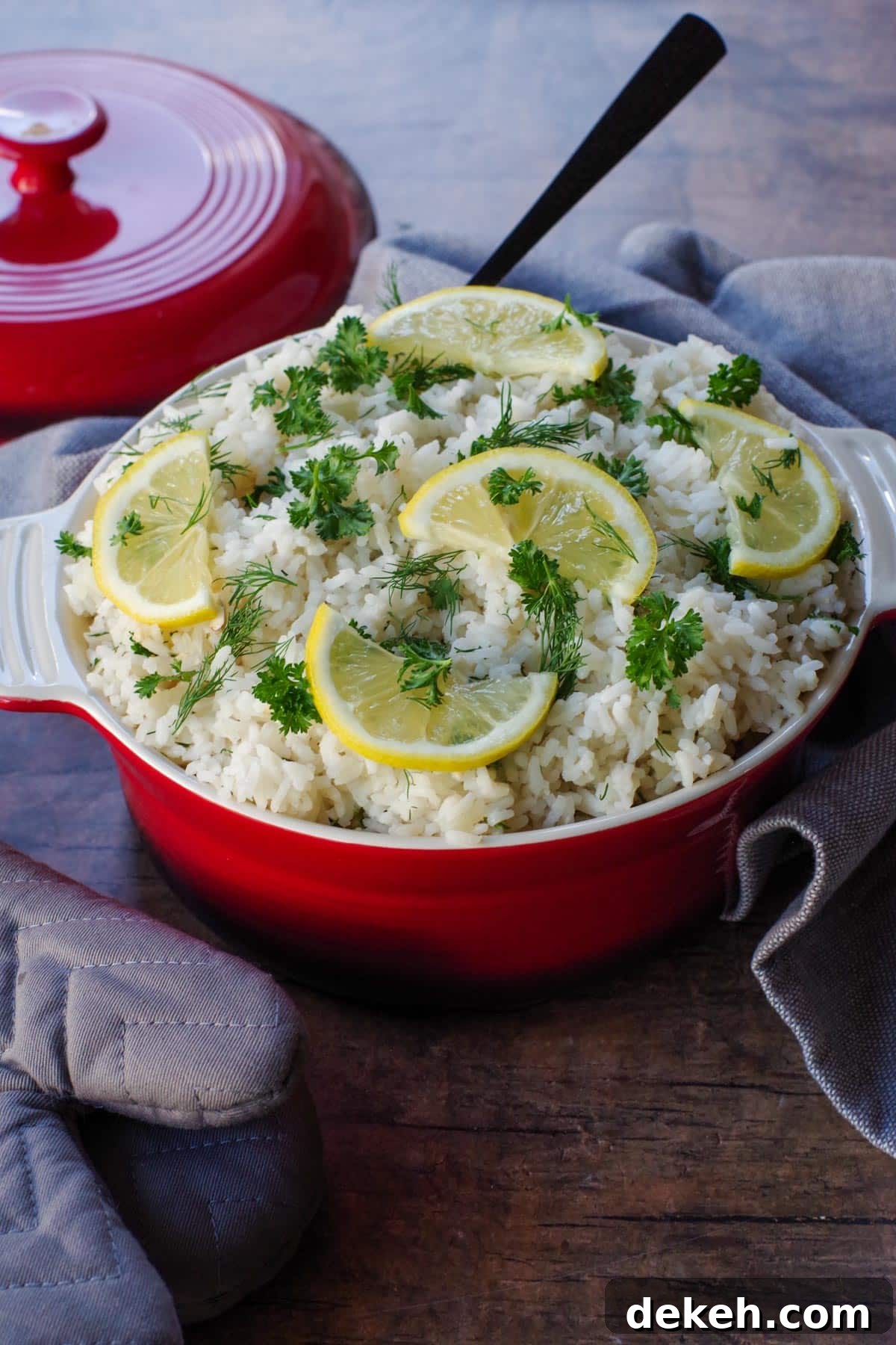 Greek rice with lemon and herb garnish in orange dish with orange lid in the background and grey oven mitt on the left side