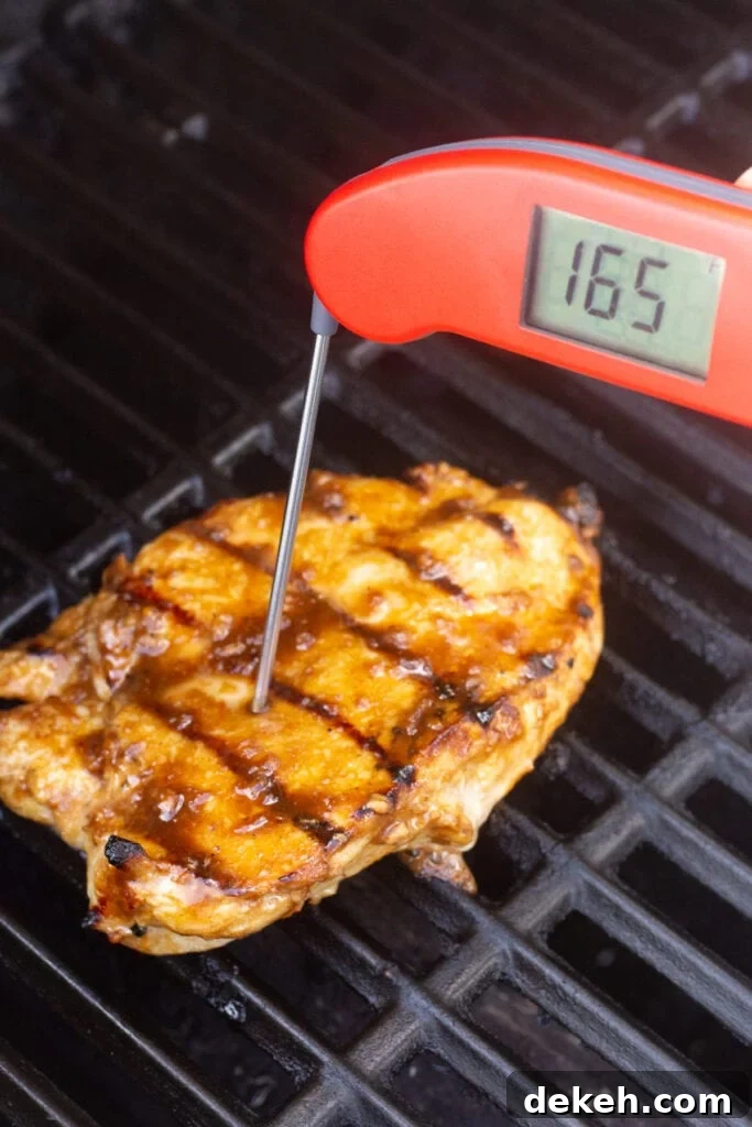A red meat thermometer checking the temperature of a grilled chicken breast on the grill, measuring 165 degrees F.