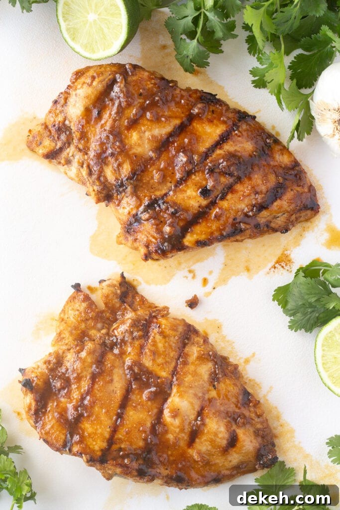 Top down shot of two grilled chicken breasts on a white cutting board with cilantro and limes surrounding them.