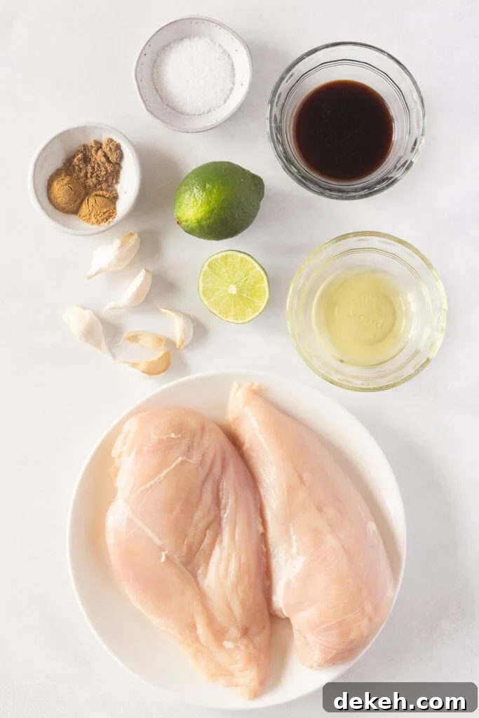 Top down shot of ingredients for grilled Mexican chicken breasts on a white background, including 2 raw chicken breasts on a white plate, 5 garlic cloves, one and a half limes, and bowls with spices and oils in them.