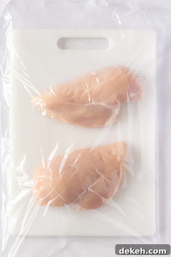 Top down shot of two raw chicken breasts on a white cutting board covered with plastic wrap.
