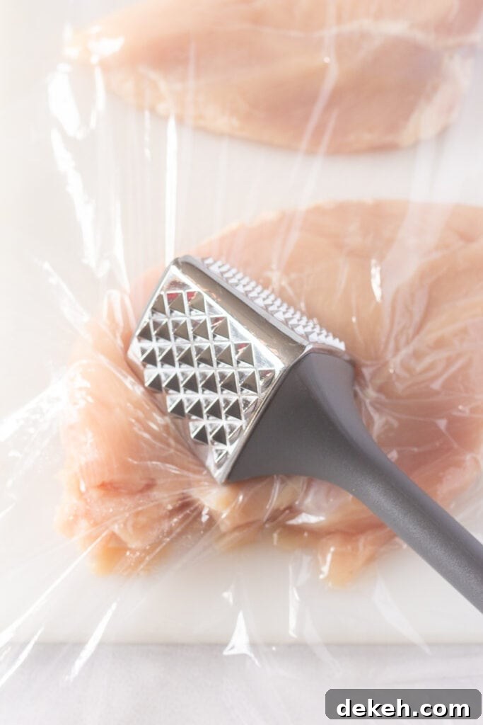 Close up of a meat mallet pounding a raw chicken breast on a white cutting board with a piece of plastic wrap on it.