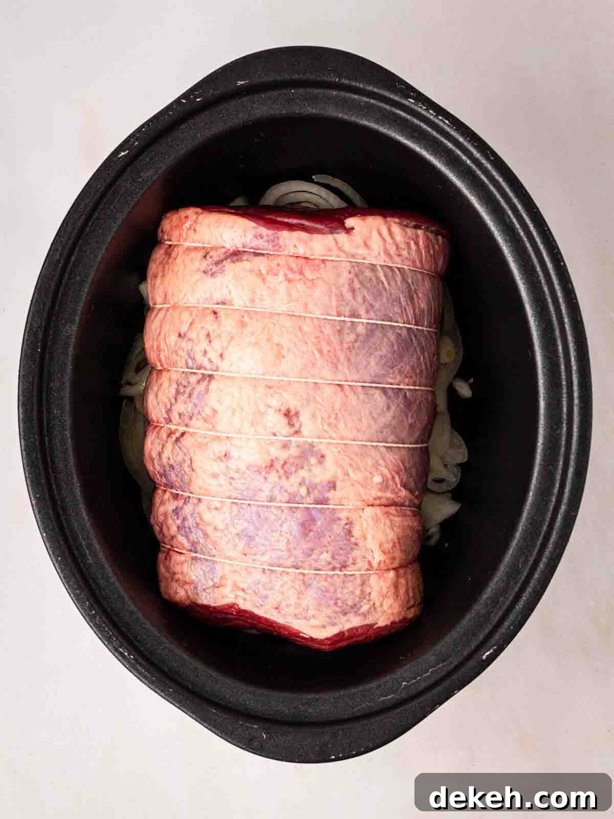 A piece of beef brisket laid on a bed of sliced onions in a large black slow cooker.
