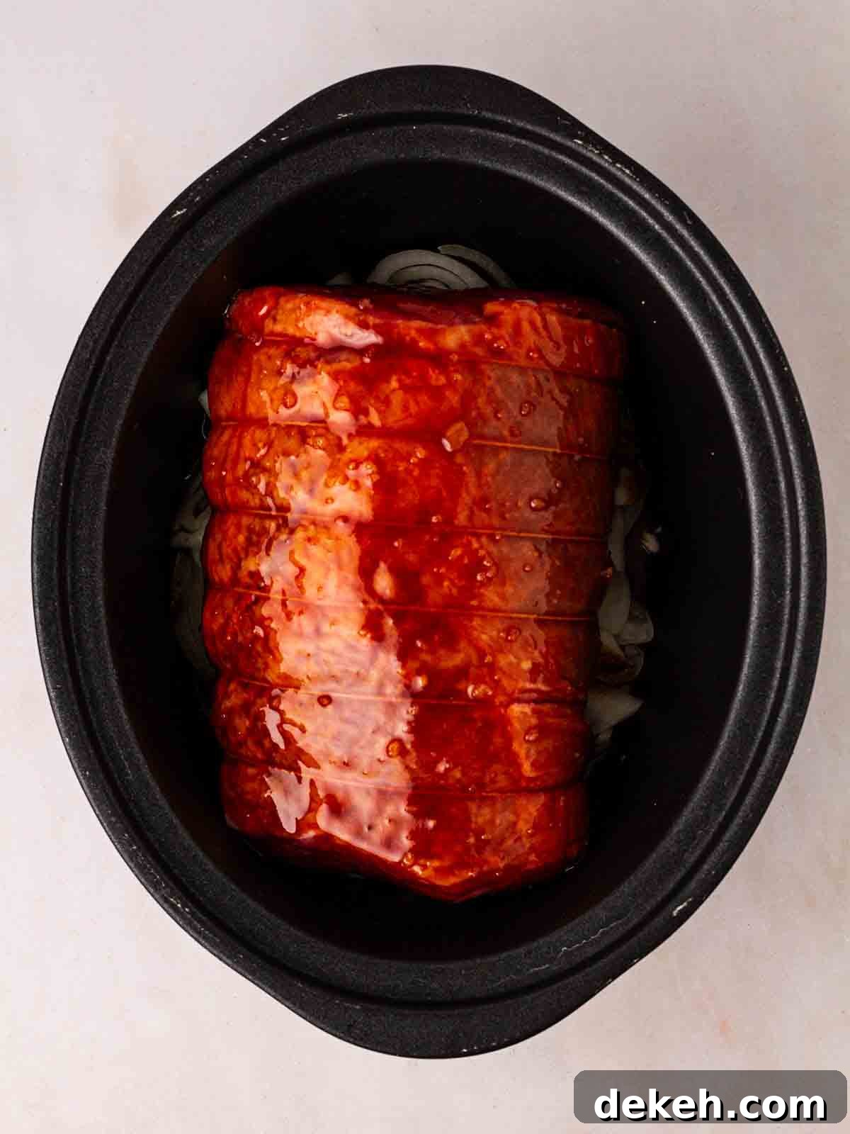 A piece of raw beef brisket in a slow cooker, topped with barbecue sauce, ready to be cooked.