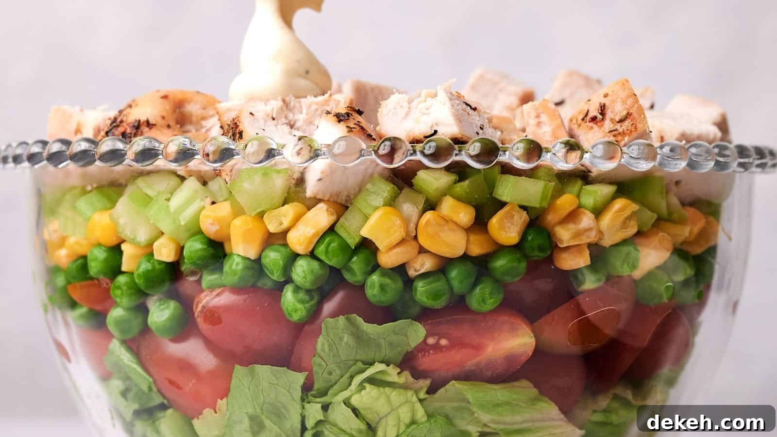A clear glass bowl layered with lettuce, cherry tomatoes, green peas, corn, diced celery, and topped with chunks of grilled chicken, garnished with a dollop of mayonnaise.
