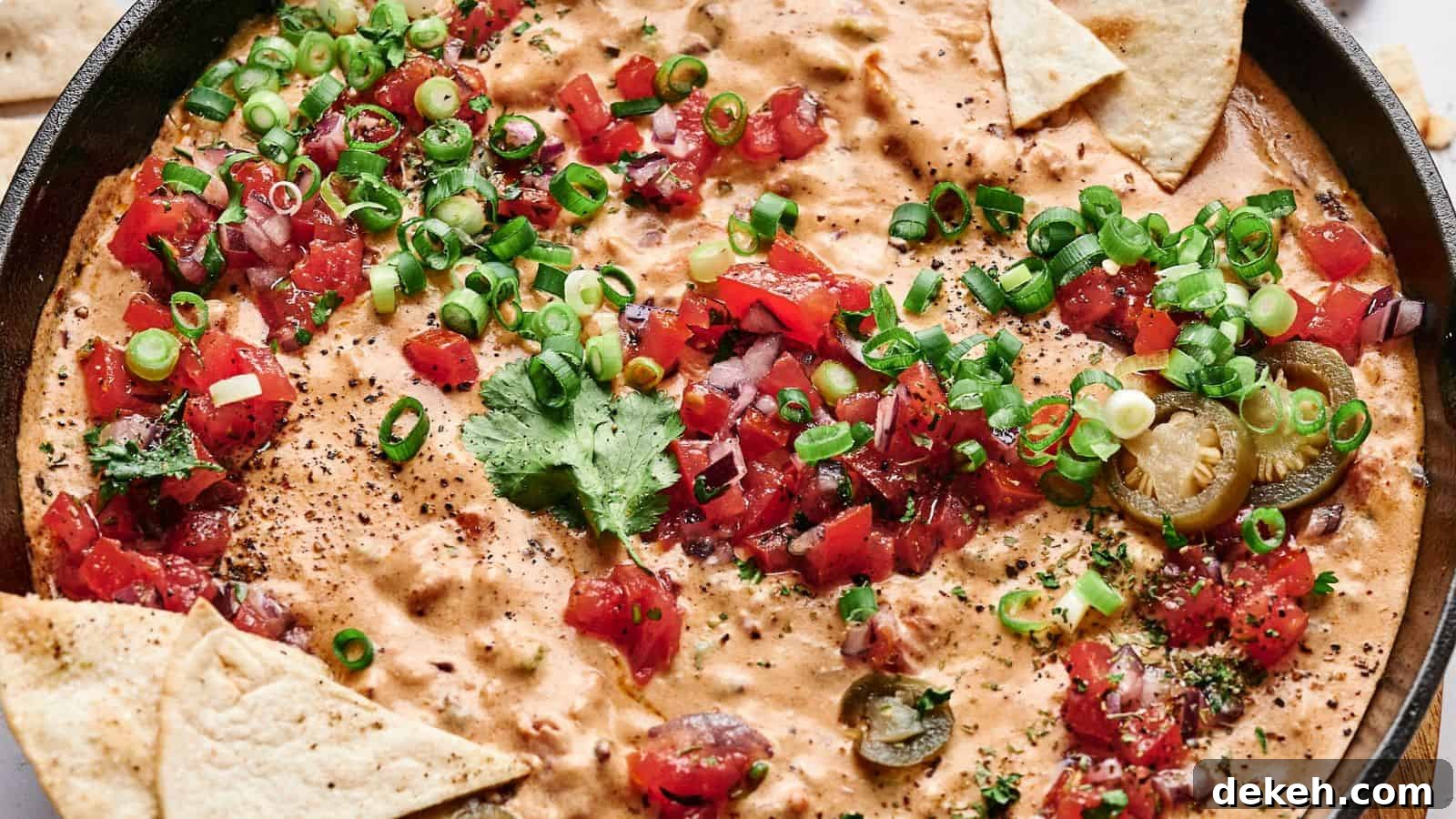A skillet filled with creamy cheese dip topped with chopped tomatoes, green onions, cilantro, and sliced jalapeños, surrounded by triangular tortilla chips.