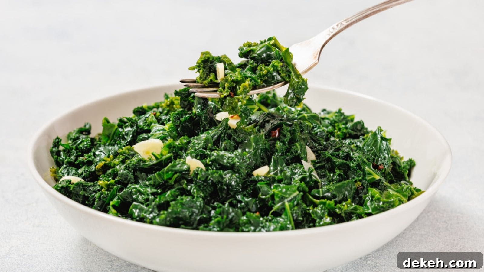A white bowl filled with sautéed kale and sliced garlic, with a fork lifting a portion from the bowl.
