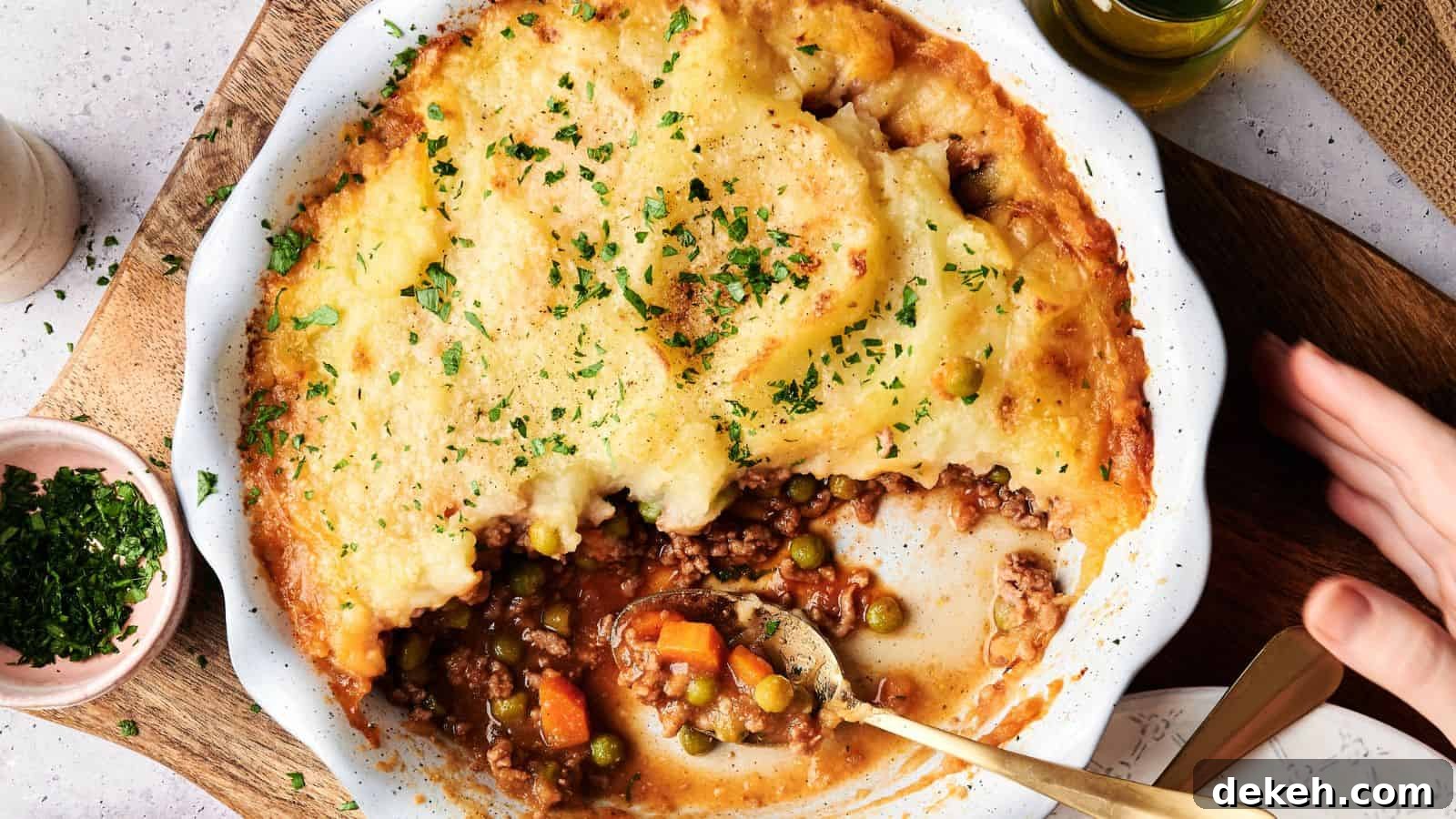 A baked shepherd's pie with a golden mashed potato topping, partially served to reveal ground meat, peas, carrots, and gravy in a white dish.