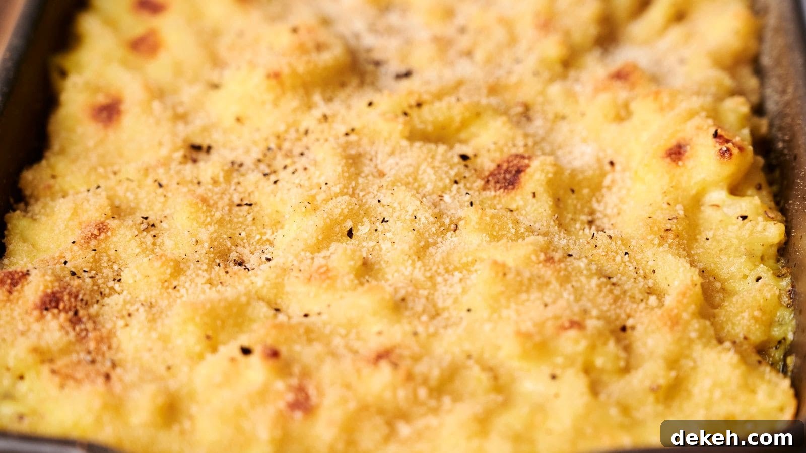 Close-up of a creamy, golden-brown macaroni and cheese dish with a crunchy breadcrumb topping reminiscent of comforting funeral potatoes. A spoon is partially submerged in the cheese sauce.