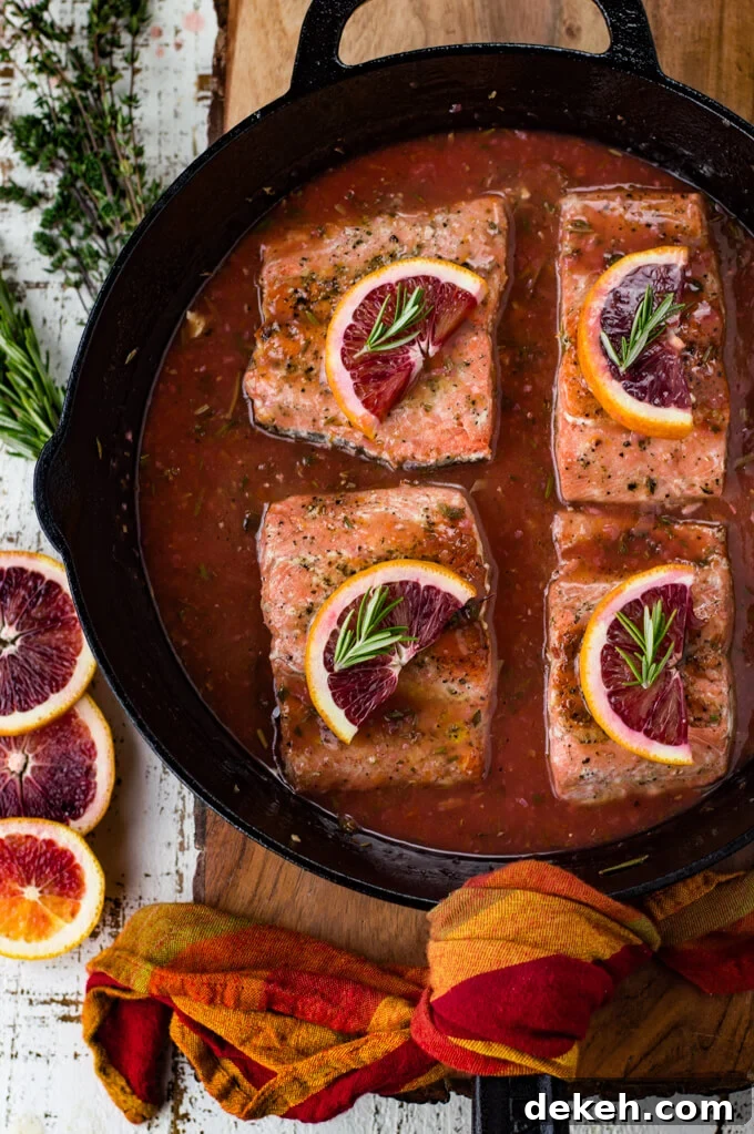 Black cast iron skillet with pieces of salmon sitting in a blood orange sauce. Each salmon has a half slice of blood orange and a small sprig of rosemary on top. Slices of blood oranges and herbs sit next to the pan.