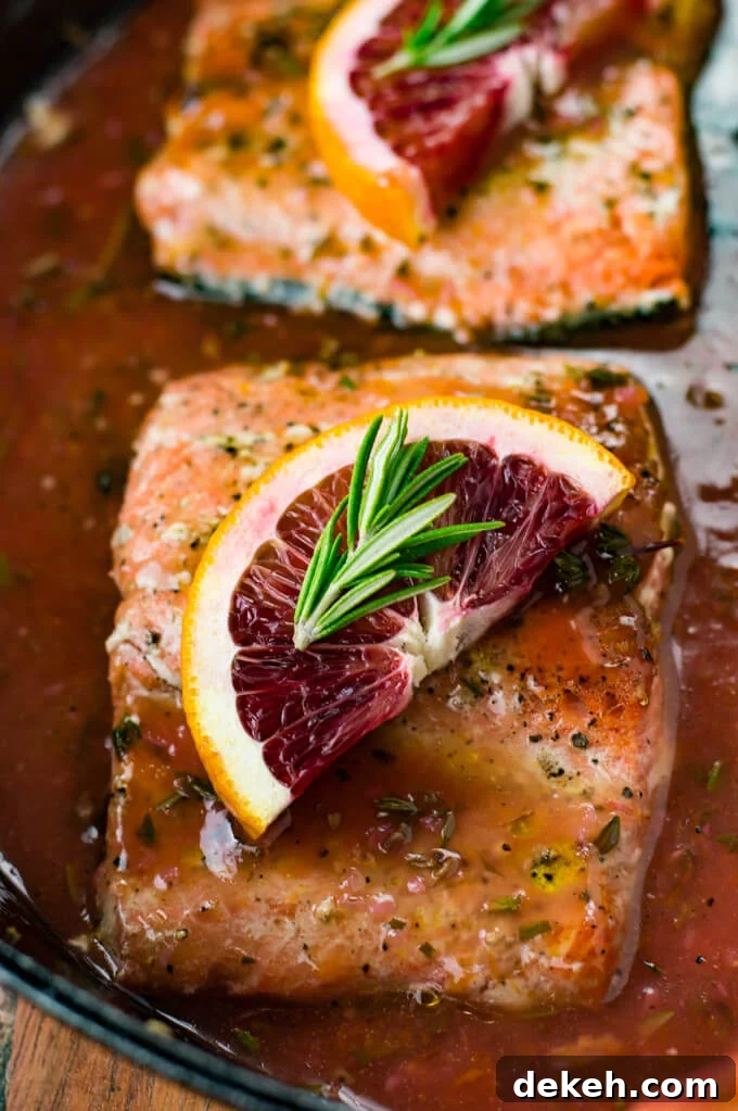 A close-up view of a piece of pan seared salmon in a blood orange sauce. A half slice of blood orange and a small sprig of rosemary sit on top.