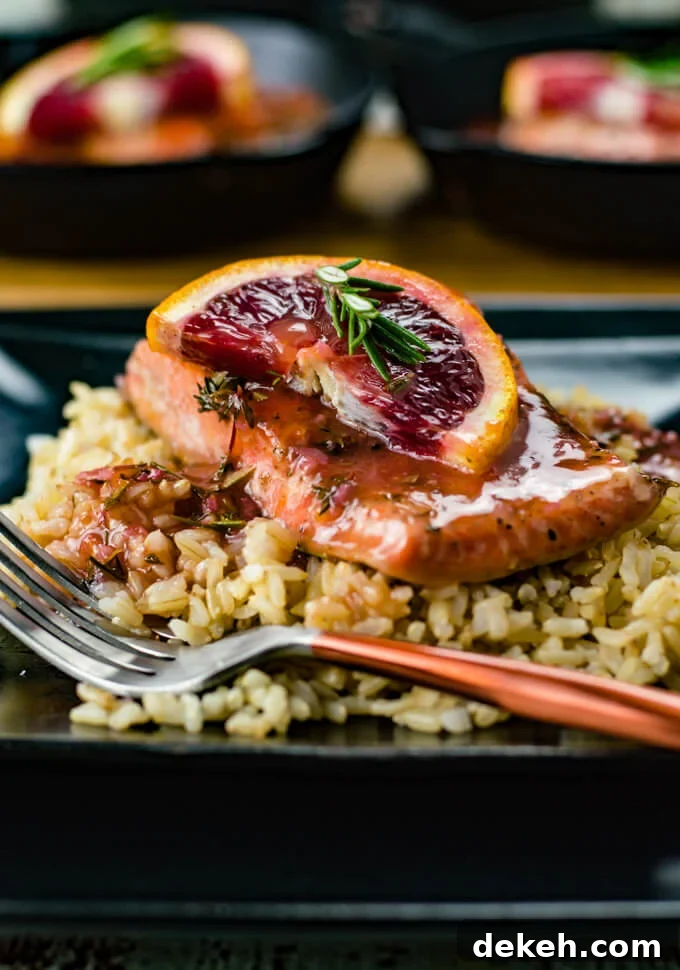 A piece of salmon with blood orange sauce sits atop a bed of rice. A half slice of blood orange with a small sprig of rosemary rests on the salmon. Two salmon pieces are blurred in the background.