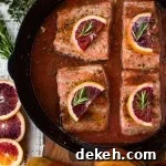 Black cast iron skillet with pieces of salmon sitting in a blood orange sauce. Each salmon has a half slice of blood orange and a small sprig of rosemary on top. Slices of blood oranges and herbs sit next to the pan.