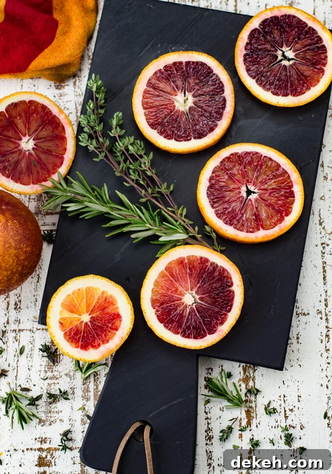 Five slices of blood oranges sit on a black cutting board with sprigs of thyme and rosemary next to them. A red and orange napkin and two halves of a blood orange sit next to the cutting board.