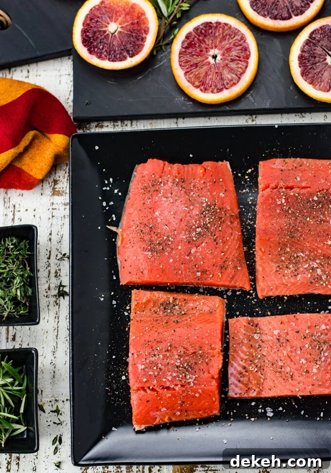 Four pieces of salmon sprinkled with salt and pepper sit on a black plate. Slices of blood oranges sit on a black cutting board above the salmon in the photo. A red and orange napkin and 2 small black bowls with thyme and rosemary sit next to the salmon.