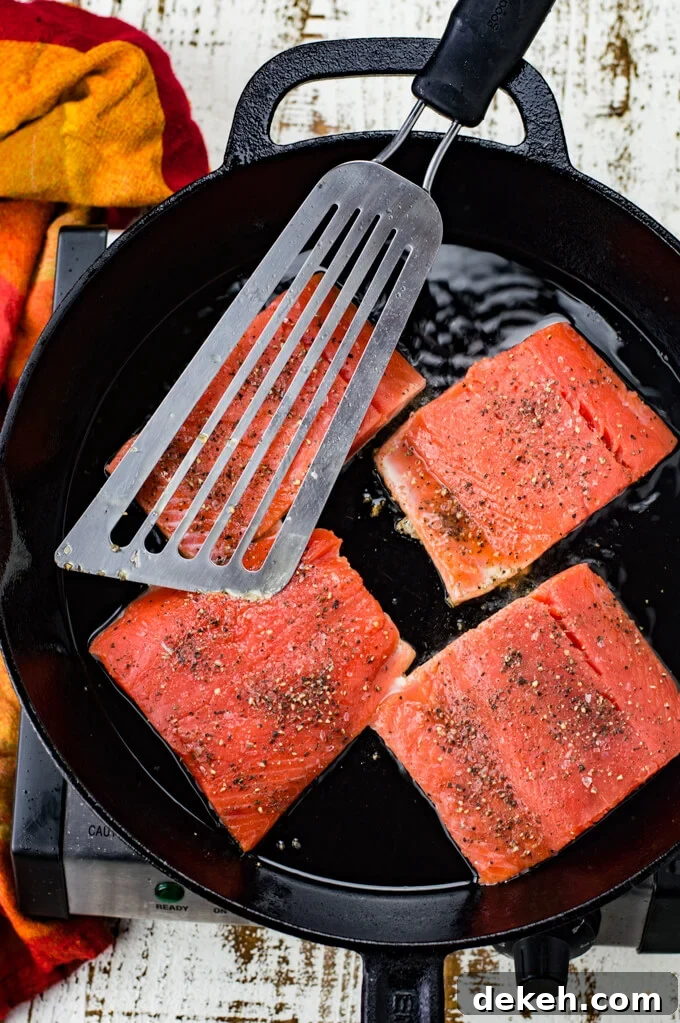 Four pieces of salmon are cooking in a black cast iron skillet. A fish spatula sits on top of them. A red and orange napkin is next to the skillet.