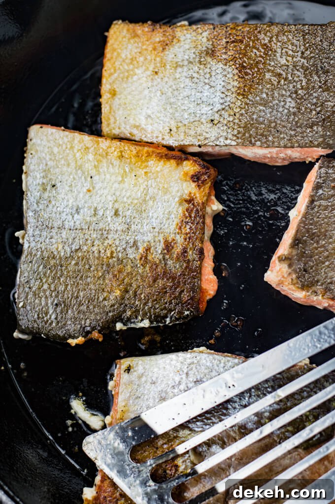 Four pieces of salmon are cooking in a black cast iron skillet with the skins up. A Fish spatula sits on top of them. 