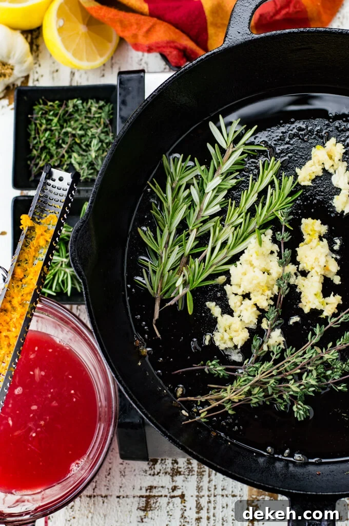 Sprigs of thyme and rosemary along with minced garlic are in a black cast iron skillet. A red and orange napkin, small bowls of thyme and rosemary, a bowl of blood orange juice sit next to the skillet. A zester with blood orange zest rests on top of the bowl.