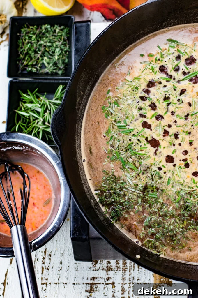 Liquid for blood orange sauce boils slowly in a black cast iron skillet. Chopped thyme and rosemary are sprinkled on top. A red and orange napkin, lemon halves, more herbs and a corn starch mixture sits next to the skillet.