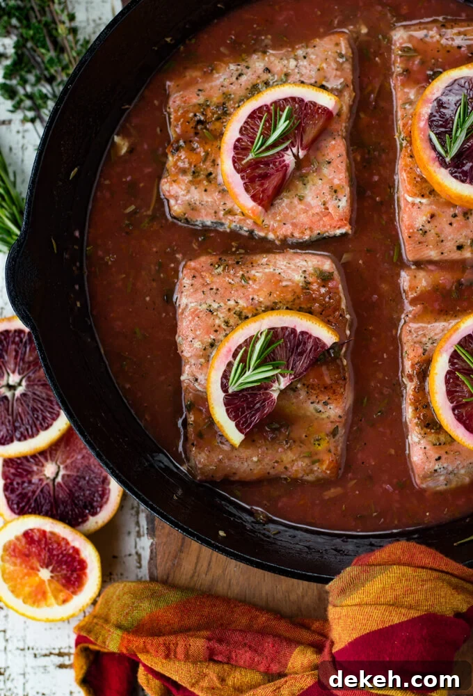 Black cast iron skillet with pieces of salmon sitting in a blood orange sauce. Each salmon has a half slice of blood orange and a small sprig of rosemary on top. Slices of blood oranges and herbs sit next to the pan.