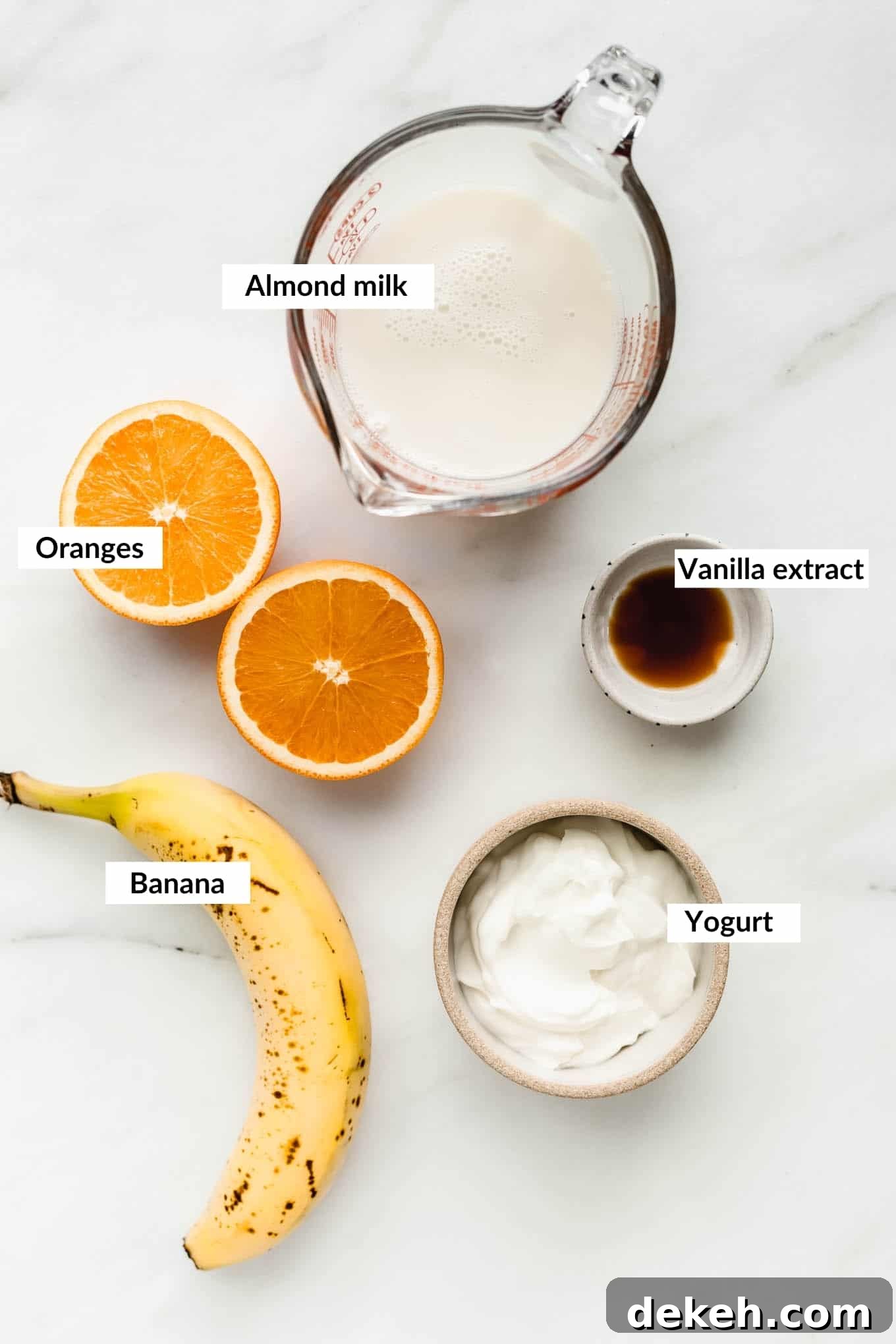 Almond milk, Orange, banana, yogurt and vanilla laid out on a white board