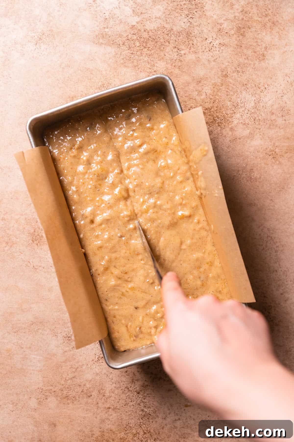 A hand dragging a butter knife through the center of the batter in a loaf pan.
