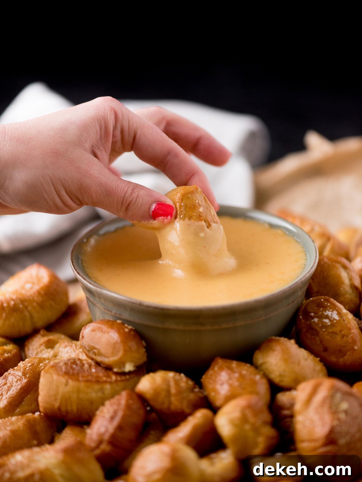 pretzel bites being dipped into beer cheese dip in a bowl. It is surrounded by pretzel bites on parchment paper
