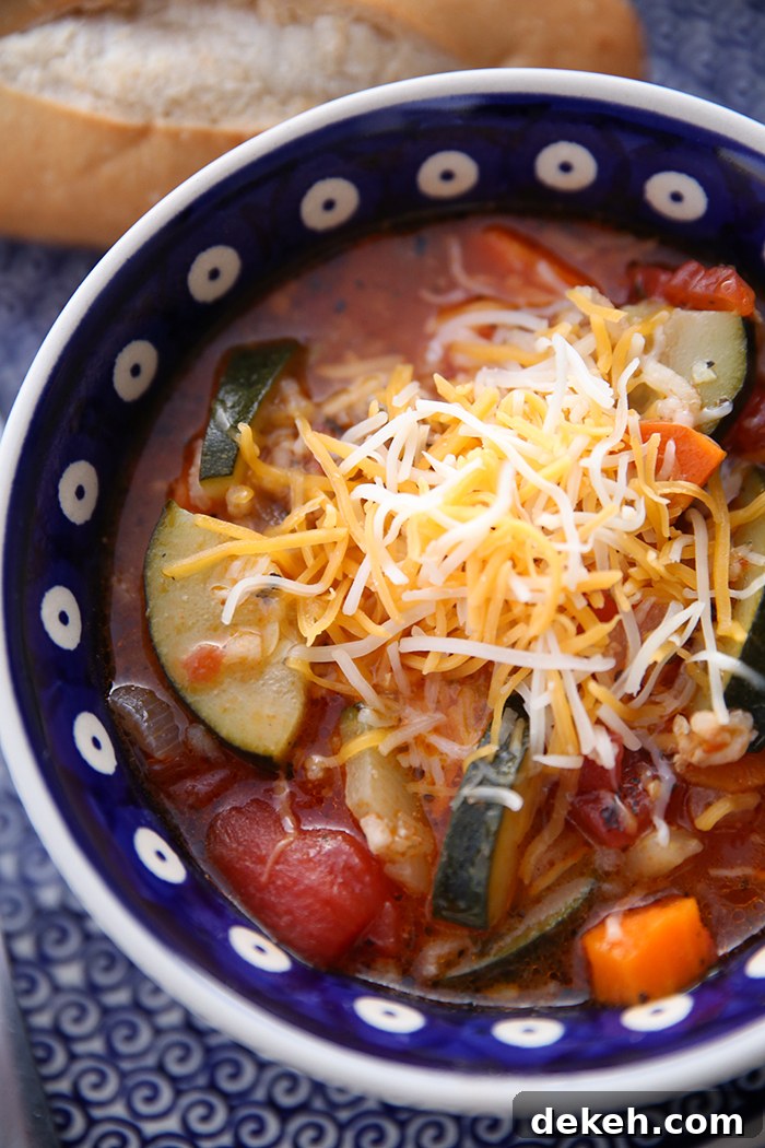 A dark blue Polish pottery soup bowl with white polka dots with a blue dot in the middle filled with stuffed zucchini soup and top with shredded colby jack cheese. At the top of the bowl is part of a crusty roll.