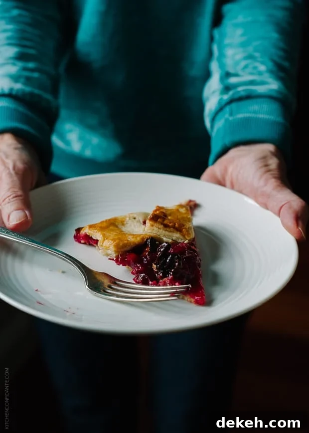 A woman in a blue sweater holding a white plate with a slice of cranberry wine galette.