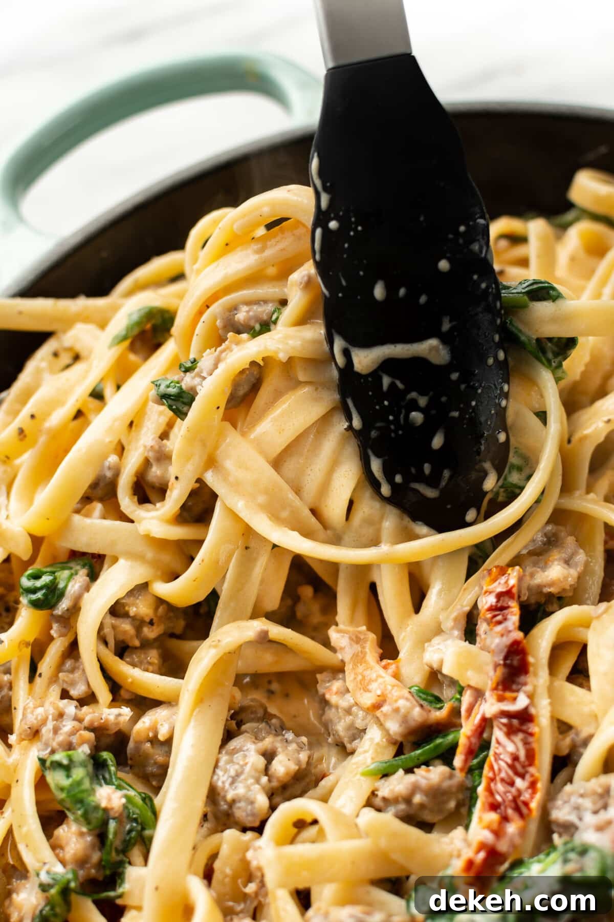 closeup of serving tongs with creamy tuscan sausage pasta in a skillet