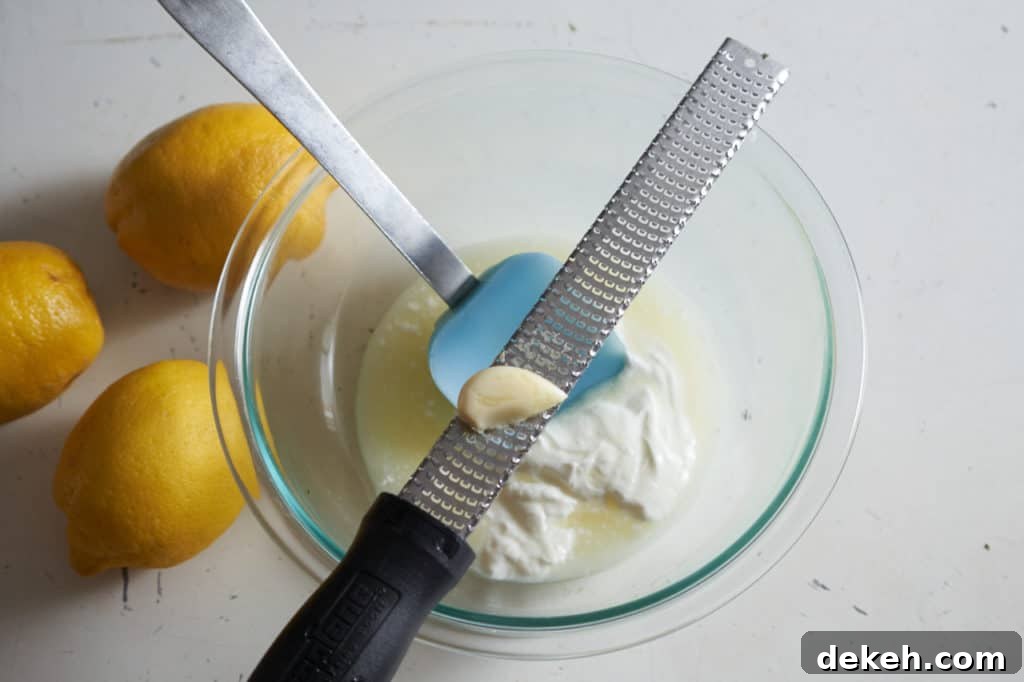 A microplane grater with a clove of garlic sits across glass bowl of yogurt and lemon juice.