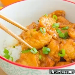Chopsticks Holding a Piece of Orange Chicken Garnished with Scallions in a Bowl with White Rice