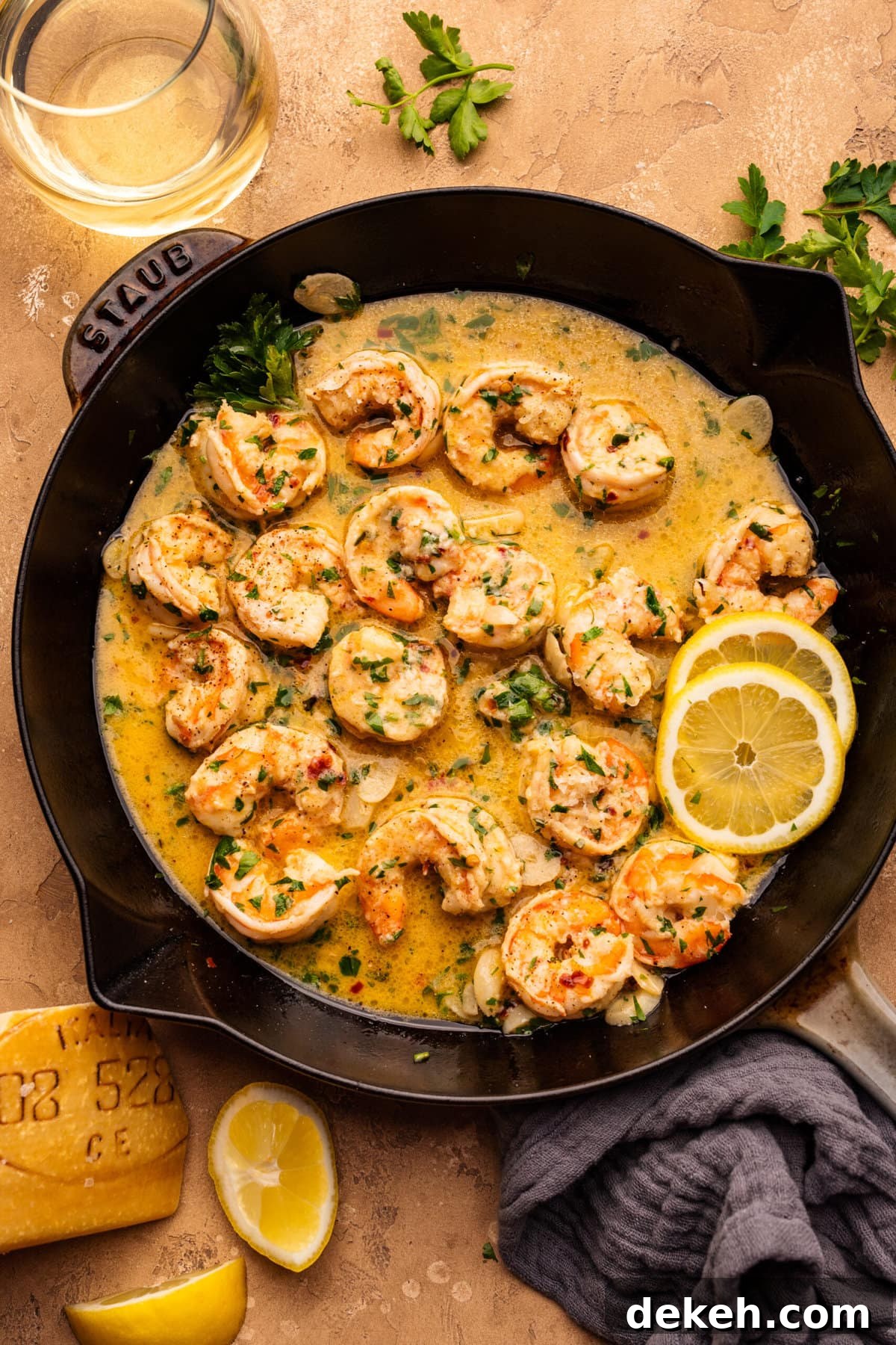 A cast iron skillet pan with shrimp scampi in it. The pan is displayed on a tan-textured background with fresh parsley, a glass of white wine, and lemon slices arranged around it.