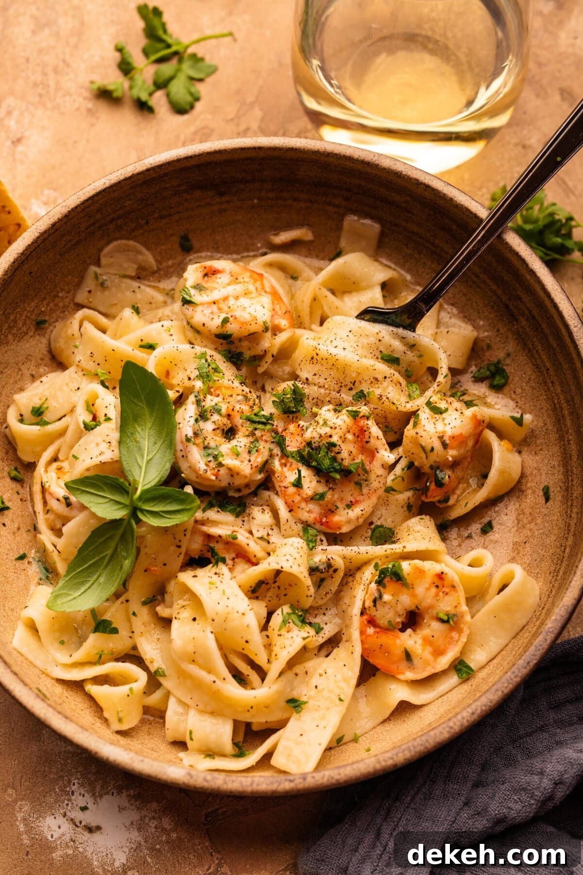 A tan bowl with shrimp scampi and tagliatelle pasta noodles in it. The dish is decorated with fresh herbs, chopped and left whole, and there is a fork in the bowl twirling the pasta. Around the bowl is a gray napkin and a glass of white wine.