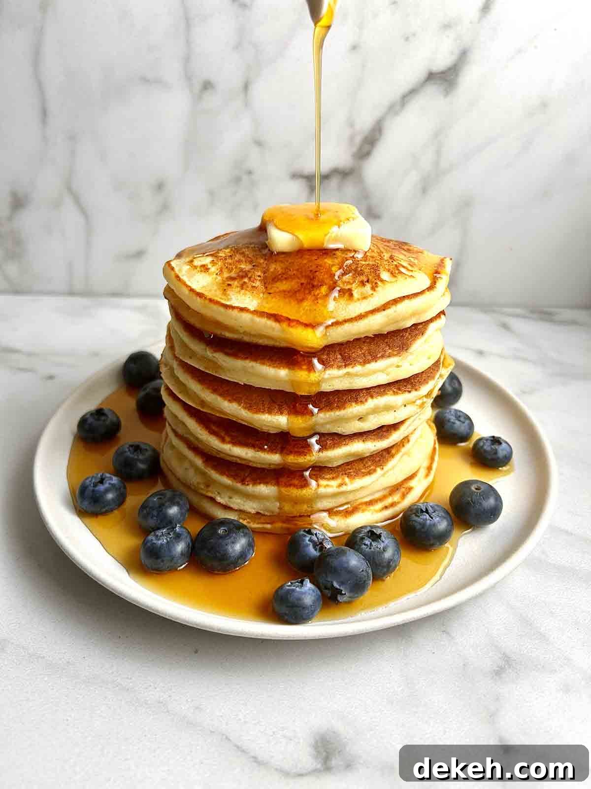 plate of ricotta pancaked stacked on top of each other; blueberries on bottom of plate, and syrup being drizzled all over.