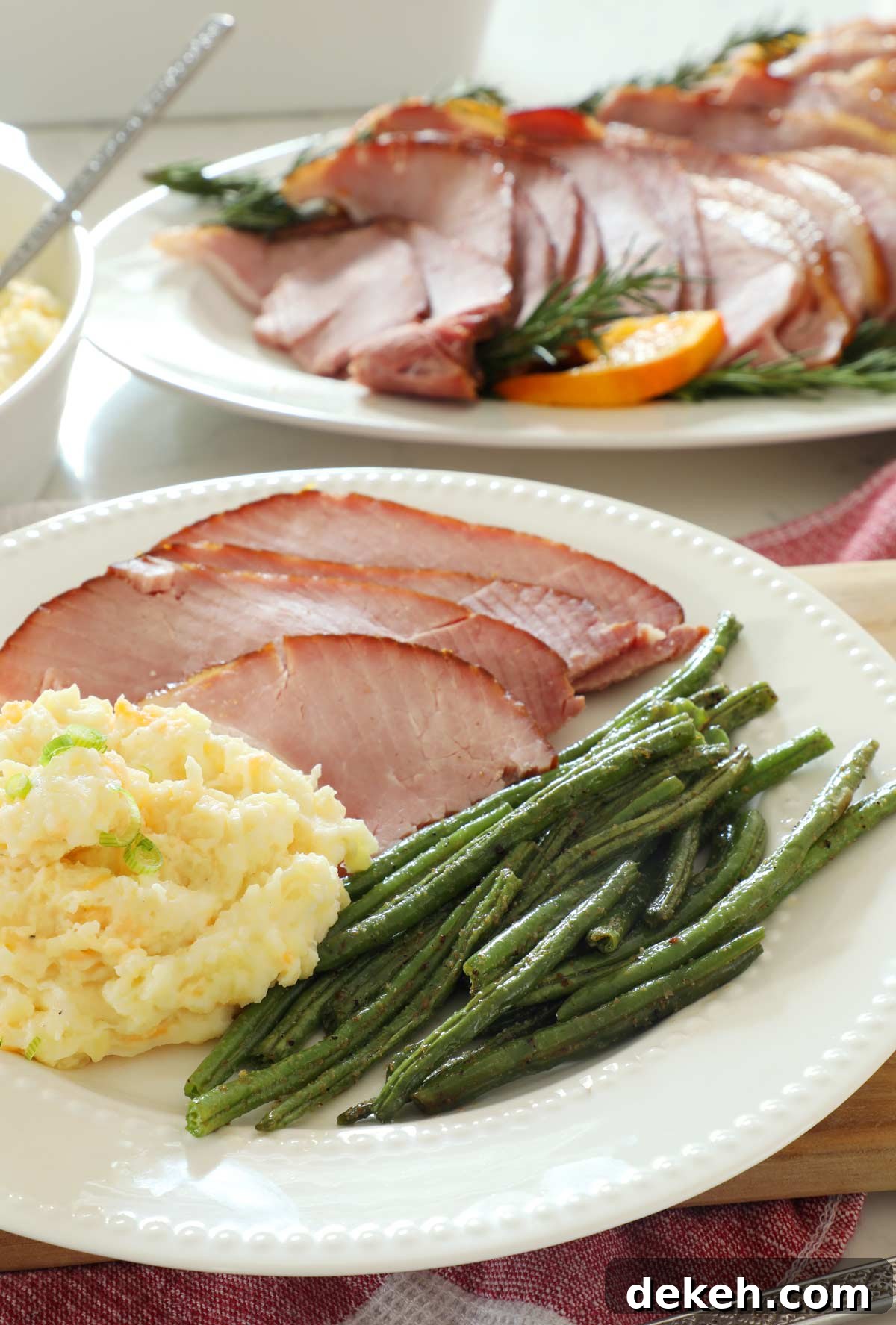 A plated ham dinner with maple glazed ham slices, cheesy mashed potatoes and baked green beans.