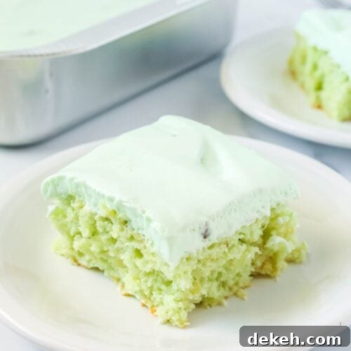 slice of pistachio pineapple cake topped with whipped cream frosting, served on a white plate. The pan of cake and another slice are in the background.